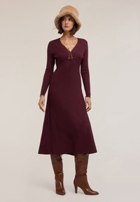 Abito a maglia a coste bordeaux con scollatura a V, maniche lunghe e vita aderente, indossato con stivali marroni con tacco alto e un cappello di pelliccia color sabbia.