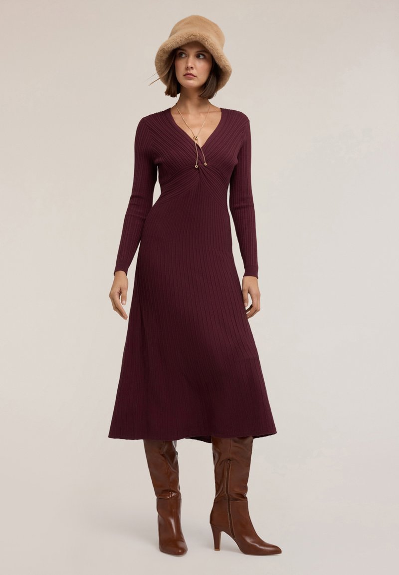 Abito a maglia a coste bordeaux con scollatura a V, maniche lunghe e vita aderente, indossato con stivali marroni con tacco alto e un cappello di pelliccia color sabbia.