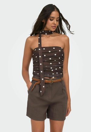 Femme portant un haut tube marron à pois blancs et une écharpe assortie, une ceinture tressée marron et un short marron taille haute, les mains dans les poches.