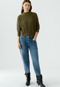 Eine Frau trägt einen olivgrünen gerippten Pullover, hochtaillierte blaue Jeans mit großen Fronttaschen und schwarze schlabberige Stiefel. Sie steht mit einer Hand in der Tasche.