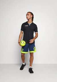 Svart sporttröja med vita detaljer och logotyp, kombinerad med marinblå shorts med neongröna sik-sak-mönster, håller en ljusgul boll.