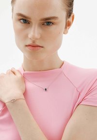 Collar de plata con un colgante de piedra oscura, llevado con una blusa rosa ajustada. También es visible una pulsera de oro rosado con corazones entrelazados.