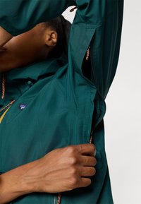 Teal windbreaker jacket met een ritssluiting, elastische aanpassingen en textuurstof met contrasterende trekkoorden.