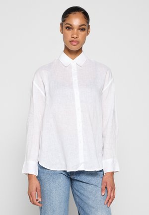Femme portant une chemise blanche ample à manches longues avec poignets et un jean bleu, debout devant un fond blanc uni.