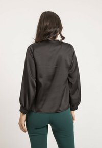 Blouse en satin noir avec une texture lisse, de longues manches bouffantes et un ourlet arrondi. Associée à un pantalon vert ajusté.