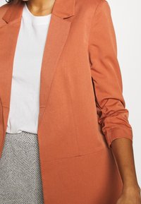 Marynarka w kolorze rdzy z usztywnionymi klapami, lekko marszczonymi rękawami i gładkim wykończeniem tkaniny. Nosi się ją na białą koszulkę i teksturowaną szarą spódnicę.