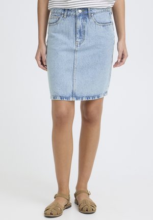 Ženska, ki nosi svetlomodro denim krilo, črtast zgornji del in rjave zaprte sandale, stoji ob belem ozadju.