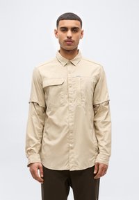 Άνδρας που κοιτάει μπροστά, φορώντας μια μπεζ πουκάμισο με μακριά μανίκια και τσέπες στο στήθος, και σκούρο παντελόνι, πάνω σε απλό ανοιχτό φόντο.