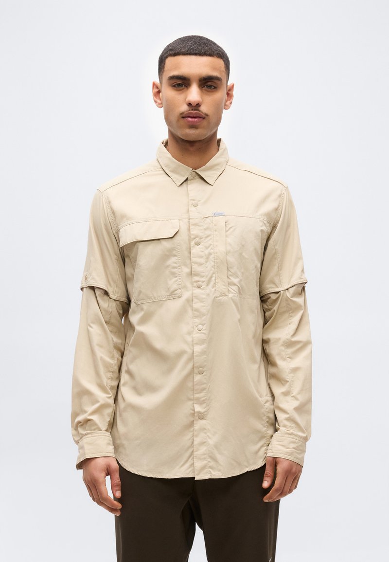 Άνδρας που κοιτάει μπροστά, φορώντας μια μπεζ πουκάμισο με μακριά μανίκια και τσέπες στο στήθος, και σκούρο παντελόνι, πάνω σε απλό ανοιχτό φόντο.