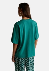 Tyrkysové bavlněné tričko s volným střihem, krátkými rukávy a kulatým výstřihem, které je spojeno s vzorovanými zelenými kalhotami.