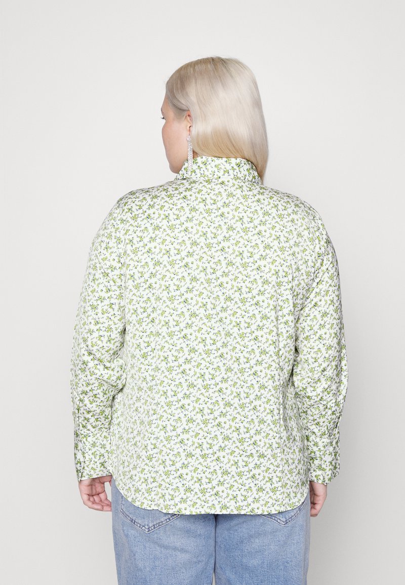 Persona bionda che indossa una camicia bianca con una piccola stampa floreale verde e jeans azzurri chiari, di spalle contro uno sfondo neutro.
