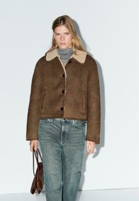 Bruine schapenvachtjas met een crèmekleurige kraag, zwarte knopen en een cropped ontwerp, gecombineerd met lichtblauwe jeans en een bruine tas.