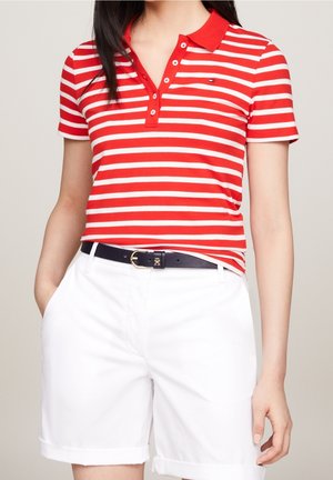 Kobieta ubrana w czerwoną i białą koszulkę polo w paski, białe szorty z wysokim stanem i czarny pasek z złotą klamrą, z rękami w kieszeniach.