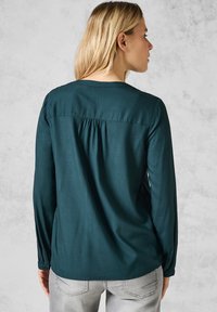 Blusa verde scuro con maniche lunghe, dettagli arricciati sul retro e polsini con bottoni. Realizzata in tessuto morbido con una leggera lucentezza.