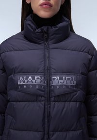 Marinblå puffjacka med hög krage. Har en dragkedjestängning och en stor logotyp på bröstet. Tillverkad av ett slätt, glänsande material med quiltad textur.