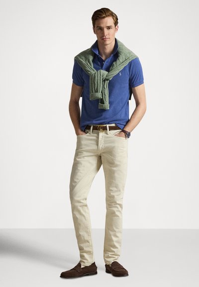 Man wearing blue polo shirt, cream pants, brown loafers, and green sweater draped over shoulders, standing with hands in pockets.