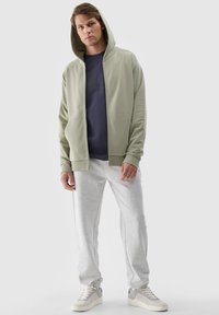Felpa con cappuccio e zip di colore verde chiaro, abbinata a una t-shirt navy e pantaloni della tuta grigi chiari. Presenta tasche e un tessuto testurizzato.