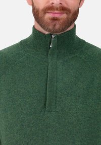 Grüner Pullover mit Reißverschluss, hohem Kragen, gerippter Struktur und mittellangem Reißverschluss. Das Material wirkt weich und gestrickt mit subtilen Farbvariationen.