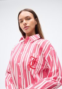 Πουκάμισο με κουμπιά και κάθετες ρίγες σε κόκκινο και λευκό χρώμα, με τσέπη στο στήθος που διαθέτει κεντημένο γράμμα και κλασικό γιακά.