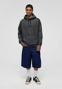 Zwarte hoodie met een voorzak, gecombineerd met blauwe denim shorts. Model draagt witte sokken en zwarte sneakers. Eenvoudig ontwerp.
