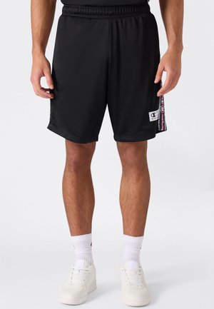 Mann trägt schwarze Champion Sportshorts mit Logo am Bein, weiße Socken und weiße Sneaker vor weißem Hintergrund.