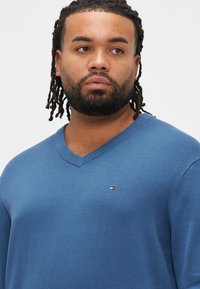 Niebieski sweter z dekoltem w kształcie litery V, wykonany z miękkiego materiału, z małym logo na lewej piersi oraz prążkowanymi wykończeniami na kołnierzu i rękawach.
