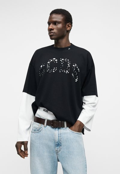 Νέος άνδρας που φοράει μαύρο υπερμεγέθες μπλουζάκι με τρύπες που σχηματίζουν το γράμμα "GCDS", λευκά μακρυμάνικα από μέσα, ανοιχτόχρωμο τζιν και καφέ ζώνη.