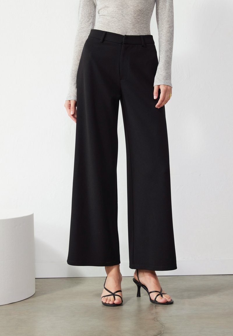 Un pantalon noir à jambes larges avec une texture lisse, une coupe ajustée et des plis nets. Associé à des talons noirs à brides.