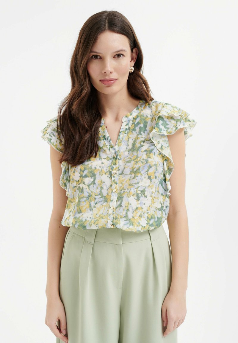 Bloemachtige blouse met groene, gele en witte patronen, volants mouwen en een knoopsluiting aan de voorkant. Gecombineerd met geplooide lichtgroene broeken.