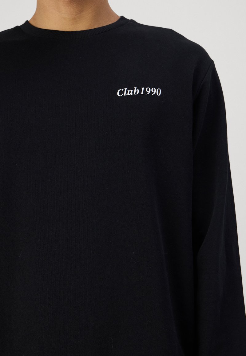 Camisa negra de manga larga hecha de algodón suave, con un pequeño logo blanco "Club1990" en el área del pecho. Diseño clásico de cuello redondo.