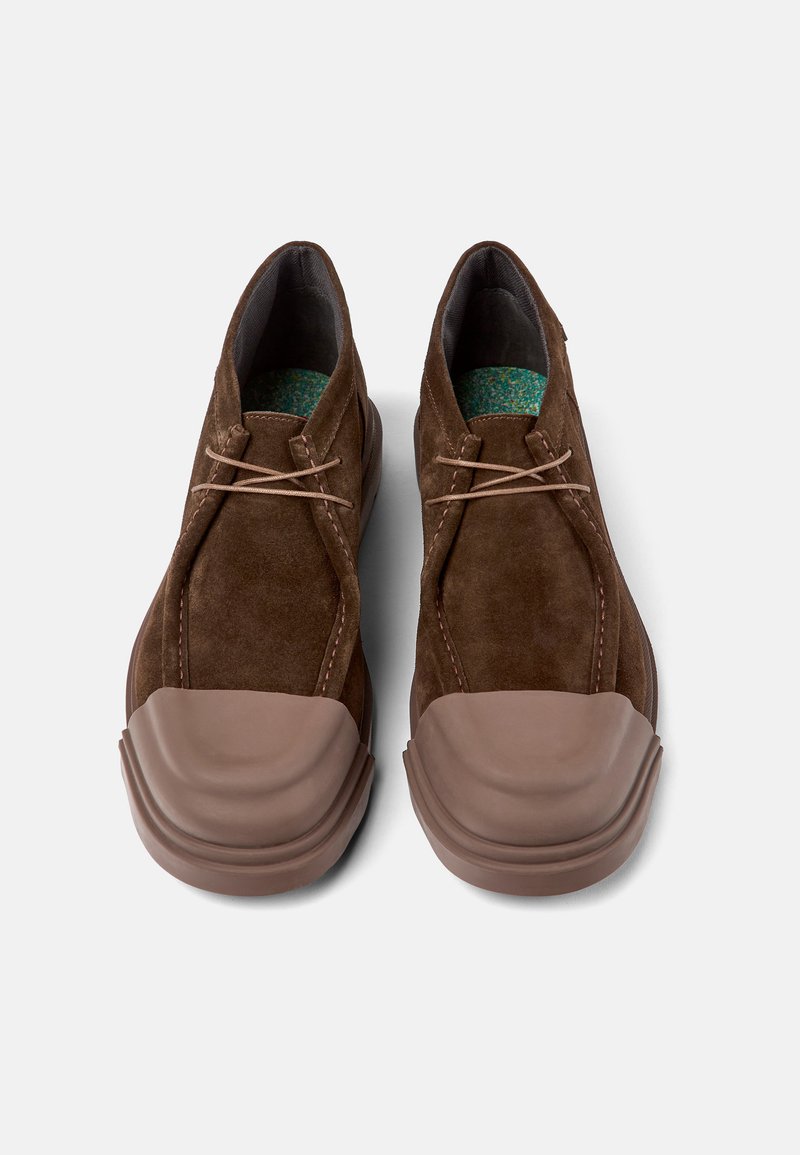 Braune Wildlederschuhe mit dicken beigen Gummikappen und -sohlen, hellbraunen Schnürsenkeln und innen sichtbaren, grün gemusterten Innensohlen.