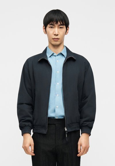 Jeune homme portant une veste noire zippée sur une chemise bleue clair boutonnée et un pantalon noir, debout devant un fond blanc.