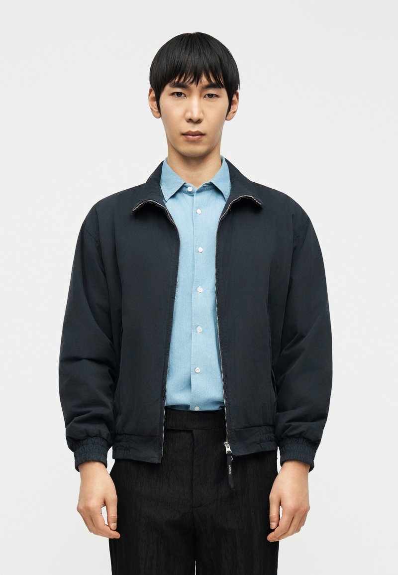 Νεαρός άνδρας που φοράει μαύρο μπουφάν με φερμουάρ πάνω από ένα ανοιχτό μπλε πουκάμισο με κουμπιά και μαύρο παντελόνι, στέκεται μπροστά σε λευκό φόντο.