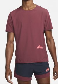 Camiseta deportiva de manga corta en color marrón, hecha de una tela ligera, con un logo de Nike Trail en rosa en la parte inferior izquierda.