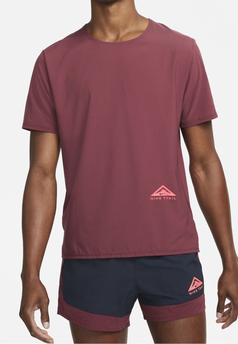 Camiseta deportiva de manga corta en color marrón, hecha de una tela ligera, con un logo de Nike Trail en rosa en la parte inferior izquierda.