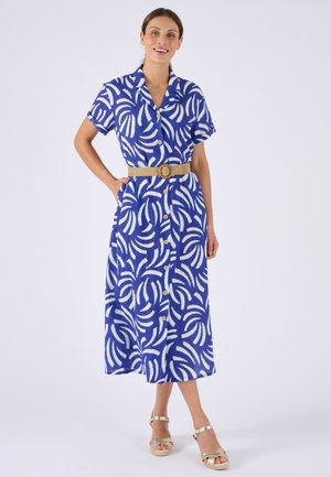 Mujer de pie con un vestido midi con botones, de estampado azul y blanco, con un cinturón beige y sandalias cuña beige.