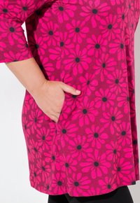 Fuchsia tunika med sort blomstermønster, har en sidelomme, rund halsudskæring og trekvartærmer, lavet af blødt stof.