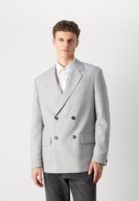 Grey plaid double-breasted blazer with two black buttons and side pockets, made of a textured fabric. Worn over a white collared shirt.