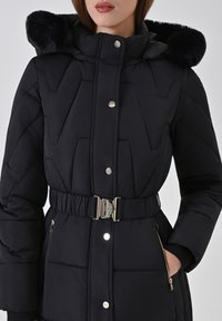 Zwarte gewatteerde jas met een capuchon van nepbont en een gestructureerd ontwerp. Voorzien van een tailleband, ritszakken en metalen sierbeslag.