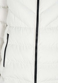 Doudoune blanche avec fermeture éclair à l'avant, motif matelassé diagonal, fermeture éclair noire et poches latérales zippées. Tissu lisse et léger.
