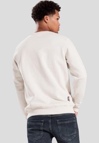 Felpa beige chiaro con maniche lunghe, polsini e orlo a coste, e dettaglio ricamato sul retro vicino al bordo inferiore. Indossata con jeans scuri.