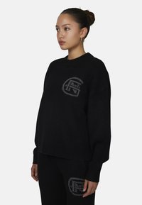 Zwarte wollen trui met een ronde hals, voorzien van een grijs logo op de linkerborst. Het ontwerp heeft een relaxte pasvorm en geribbelde boorden.