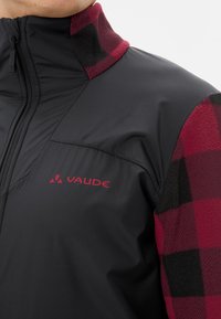 Zwarte en rode fleecejack met een ritssluiting, voorzien van een geruit patroon op de kraag en schouder, met een rood "Vaude" logo.