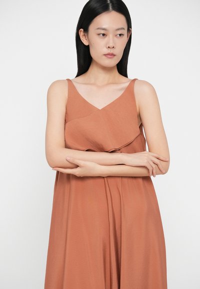 Γυναίκα με μακριά μαύρα μαλλιά, ντυμένη με ένα αμάνικο �χάλκινο φόρεμα, με σταυρωμένα τα χέρια, να κοιτάζει στο πλάι, μπροστά σε έναν μονόχρωμο λευκό τοίχο.