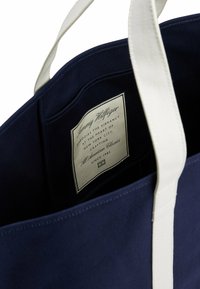 Borsa tote in tela blu navy con manici in cotone bianco e un'etichetta interna che dettaglia il marchio e le specifiche, con una texture liscia.