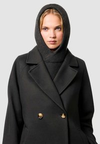 Cappotto in misto lana nero con design doppiopetto, dotato di bottoni dorati e un cappuccio in maglia scura coordinato. Texture liscia e vestibilità su misura.