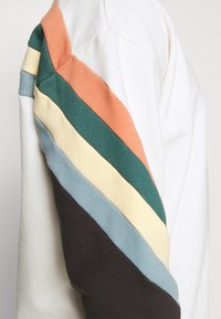 Felpa caratterizzata da un corpo bianco con strisce diagonali multicolori in arancione, verde, giallo, blu e marrone lungo una manica.