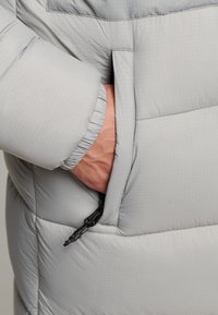 Material de chaqueta acolchada gris claro con un patrón de cuadrícula. Cuenta con un bolsillo lateral y un puño elástico. Detalle visible de tirador de cremallera resistente.