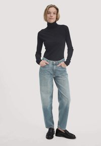 Schwarzer gerippter Rollkragenpullover, hellblaue high-waisted gerade Jeans und schwarze Slipper. Das Model steht mit den Händen in den Taschen vor einem hellen Hintergrund.