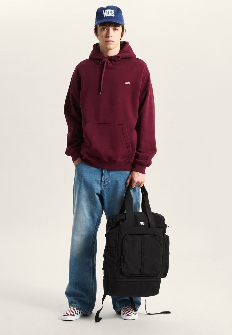 Sweat à capuche bordeaux avec poche kangourou, casquette Vans bleue, jean ample délavé clair, sac à dos noir avec plusieurs poches et sangles, slip-ons à carreaux.